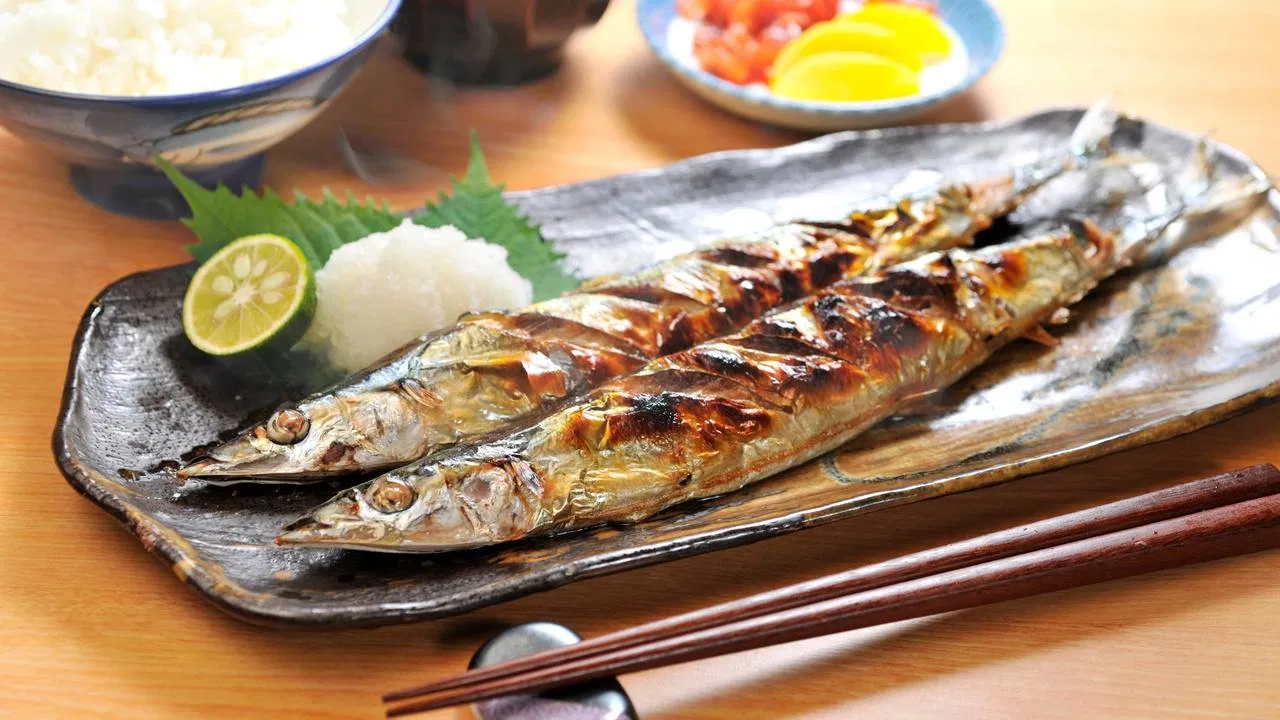 長い角皿に、皮がこんがり焼けたサンマが2尾並んでいます。手前に青じその葉、輪切りのすだち、大根おろし。奥にはごはん、お味噌汁のお椀、たくあんと漬物の小皿があり、手前には箸と箸置きが置かれた写真